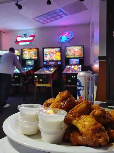 A plate of glazed wings with dipping cups and a Pabst can, pinball machines glowing behind it